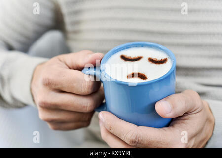 Primo piano di un giovane uomo caucasico con un blu tazza di cappuccino con una faccina triste disegnata con il cacao in polvere sulla schiuma di latte Foto Stock