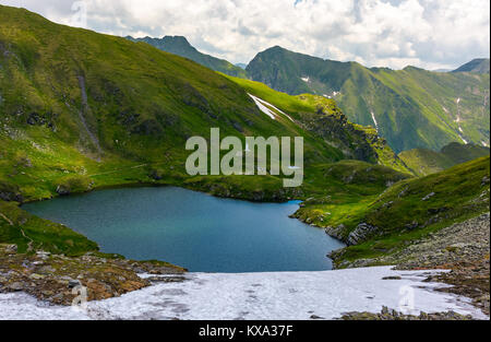 Splendida vista del lago in alta montagna. incantevole paesaggio estivo con neve sulle colline erbose Foto Stock