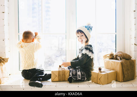 I bambini del fratello e sorella di età prescolare sedere alla finestra su un soleggiato il giorno di Natale e di giocare con le scatole dei regali avvolti in carta.Essi sono vestiti kn Foto Stock