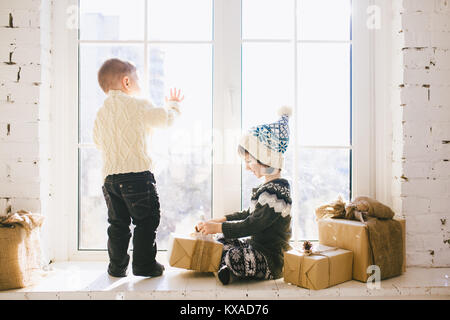 I bambini del fratello e sorella di età prescolare sedere alla finestra su un soleggiato il giorno di Natale e di giocare con le scatole dei regali avvolti in carta.Essi sono vestiti kn Foto Stock