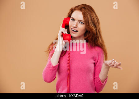 Close up ritratto di un confuso pretty redhead girl parlando sul tubo di telefono isolato su sfondo beige Foto Stock