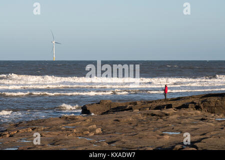 Un pescatore che pesca con asta e linea appena a nord di Seaton Sluice, con una turbina eolica in background, Northumberland, England, Regno Unito Foto Stock