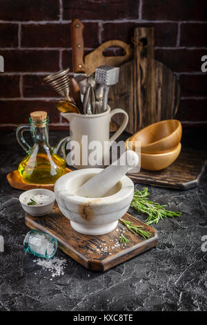 Mortaio di marmo per le spezie, sale e olio di oliva e rametti di rosmarino su un tagliere di legno Foto Stock