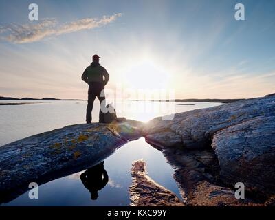 Giovane uomo in piedi con lo zaino. Escursionista sulla pietra sulla riva del mare al tramonto colorato sky. Bellissimo paesaggio con uomo sportivo rocce e mare nuvole Foto Stock