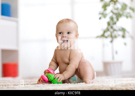 Carino il bambino nel pannolino si siede sul tappeto e gioca con il giocattolo a casa. Profondità di campo. Foto Stock
