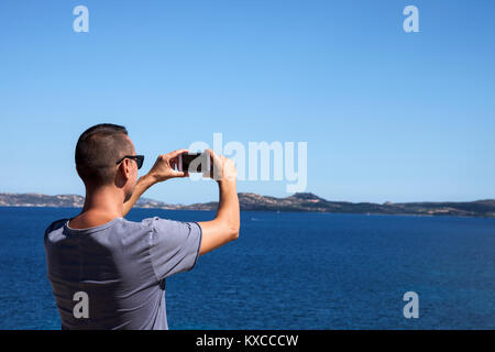 Primo piano di un giovane uomo caucasico visto da dietro di scattare una foto del mare della Costa Smeralda, in Sardegna, Italia, con il suo smartphone, con un bla Foto Stock