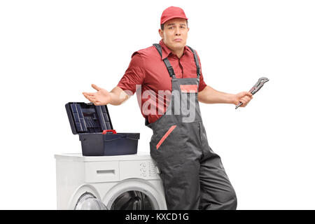 Incerto aggiustatore con una chiave appoggiata ad una macchina di lavaggio e di gesti con le mani isolati su sfondo bianco Foto Stock