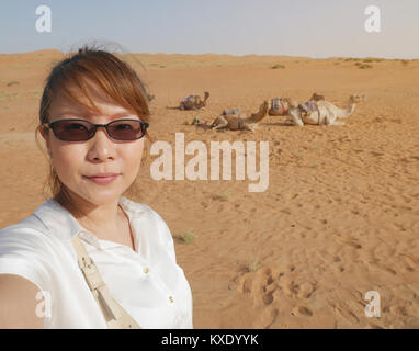 Femmina asiatica turistica prendendo selfie a Wahiba desert in Oman con sullo sfondo di un allevamento di cammelli seduti sulla sabbia calda, in attesa che la corsa in cammello attività Foto Stock