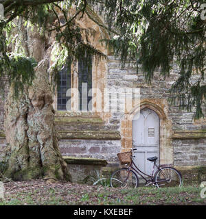 Vecchia bicicletta con cestello appoggiata contro la vecchia chiesa porta accanto ad albero nel tranquillo villaggio inglese. Foto Stock