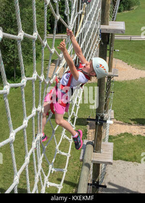 Audace giovane ragazza salendo su una fune di elevata net su un percorso ad ostacoli. Per raggiungere un appiglio, esprimendo il coraggio e determinazione. Foto Stock