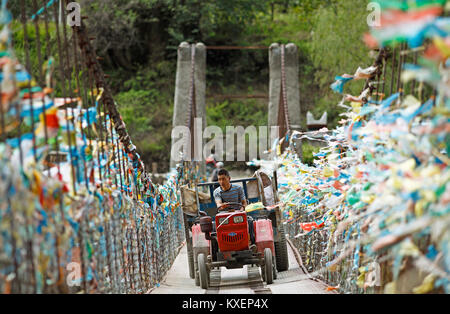 Il contadino di unità con un trattore su un ponte di sospensione decorata con bandiere di preghiera, Jiaju Village, Distretto autonomo di Garzê Foto Stock
