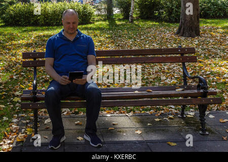 Uomo in camicia blu con lettura digitale compressa su una panchina nel parco Foto Stock