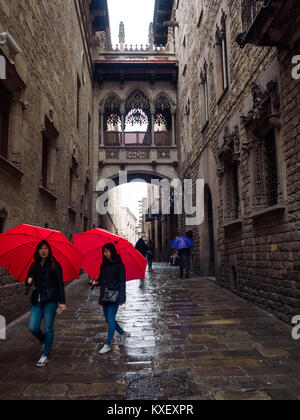 Due donne turistiche camminano attraverso il quartiere gotico di Barcellona, sotto la pioggia e l'arco in Carrer del Bisbe (via Vescovo), portando con sé lo stesso ombrello rosso Foto Stock