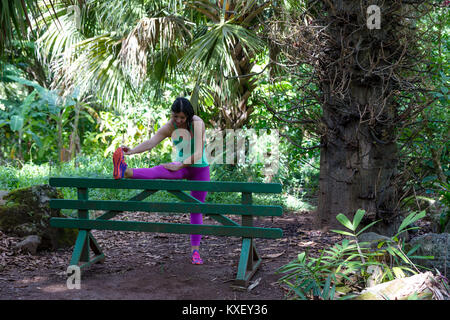 Atletica Giovane donna che lavora fuori in un lussureggiante parco verde in Oahu, Hawaii facendo stretching esercizio utilizzando una rustica panca in legno per il supporto Foto Stock