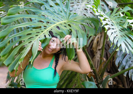 Carino donna giocoso che spuntavano da sotto una grande foglia verde di monstera deliciosa, o Delicious Monster, impianti su Oahu, Hawaii con un sorriso Foto Stock