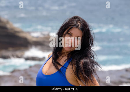 Attraente naturale battente giovane donna con lunghi capelli tousled sulle rocce al punto di cielo, Oahu, Hawaii sorridente in telecamera Foto Stock