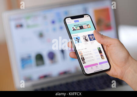 Primo piano di una donna di sfogliare musica su iTunes Store su Apple iPhone X. mano che tiene un iPhone con un laptop macbook lo sfondo. Foto Stock