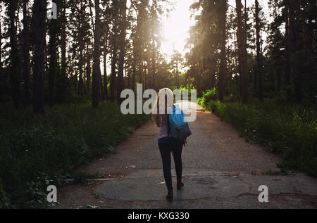 Vista posteriore della donna con zaino camminando sulla strada di campagna tra gli alberi nelle foreste Foto Stock