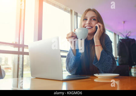 Felice giovane donna tanga con tazza di cofe parlando al telefono in presso il cafe. Computer portatile sul tavolo. Foto Stock