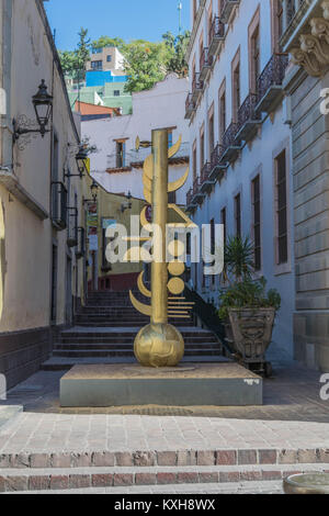 Oro statua astratta su un mattone strada asfaltata, con mattoni i passaggi e colorate case ed edifici tiered giù per una collina, in Guanajuato, Messico Foto Stock