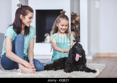 Madre sorridente che guarda la figlia spazzolando i capelli del cane in soggiorno a casa Foto Stock