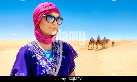 Ritratto di donna bella in arabo abbigliamento tradizionale sullo sfondo di turisti a cavallo di cammelli. Deserto del Sahara, Tunisia, Nord Africa Foto Stock