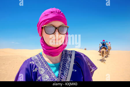 Ritratto di donna bella in arabo abbigliamento tradizionale sullo sfondo di turisti a cavallo di cammelli. Deserto del Sahara, Tunisia, Nord Africa Foto Stock
