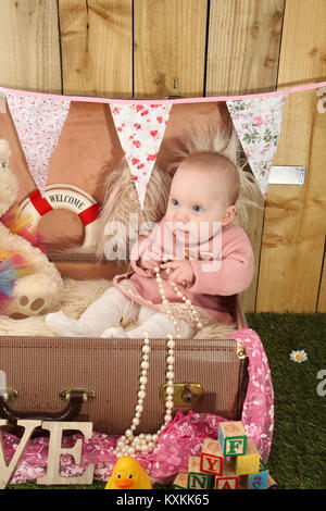 Pretty baby ragazza seduta in valigia a giocare con i giochi in giardino Foto Stock