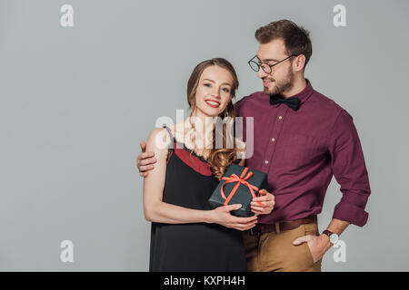 Bello elegante uomo sorridente e abbracciando il giovane e bella donna azienda confezione regalo isolato su grigio Foto Stock