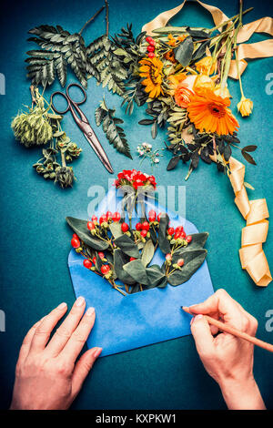 Mani femminili di scrivere con la matita sul blu aperto avvolgono con fiori. Fioraio con fiori autunnali e apparecchiature di decorazione blu su sfondo tabella, parte superiore Foto Stock