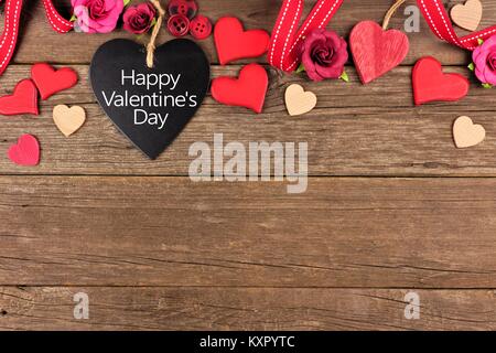 Happy Valentines Day messaggio su una a forma di cuore tag di lavagna con nastro e cuori bordo contro un rustico sfondo legno Foto Stock