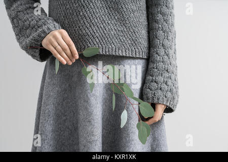 Ritratto di una ragazza in un grigio pullover a maglia ed un mantello su sfondo grigio chiaro in studio. Essa detiene un ramo di eucalipto nella mano destra Foto Stock