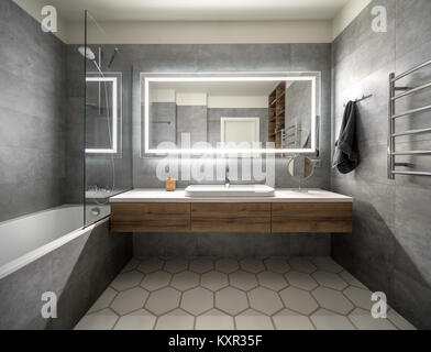 Bagno in stile moderno con tonalità grigio e piastrelle bianche. Vi è un grande specchio con lampade luminose, da tavolo con cassetti in legno e lavandino, vasca da bagno con sh Foto Stock