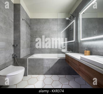 Bagno moderno con il grigio e il bianco delle piastrelle. Vi è un grande specchio con lampade luminose, da tavolo con cassetti in legno e lavandino, vasca da bagno con doccia e un Foto Stock