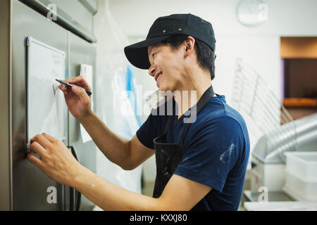 Uomo che lavora in una panetteria, indossare un cappello da baseball e grembiule, la scrittura della nota sulle piccole whiteboard, utilizzando il telefono e sorridente. Foto Stock
