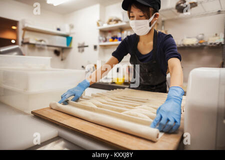 La donna a lavorare in una panetteria, indossare guanti protettivi e maschera, ponendo la pasta su grande tavola di legno. Foto Stock