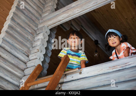 Basso angolo vista del ragazzo e ragazza con i capelli neri guardando fuori della finestra, scaletta appoggiata contro la parete di legno. Foto Stock