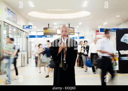 Monaco buddista con testa rasata indossa black robe in piedi all'interno di una stazione ferroviaria, tenendo mala, gente camminare passato. Foto Stock
