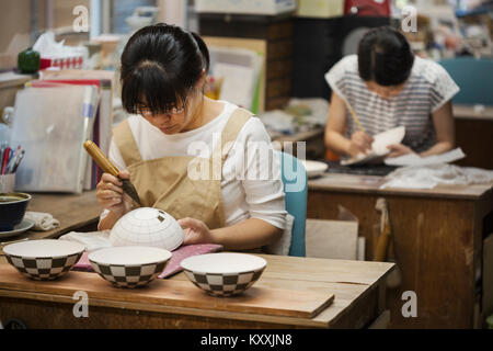 Donna che lavorano in un giapponese workshop di porcellana, pittura disegno geometrico su bianco ciotole con pennello. Foto Stock