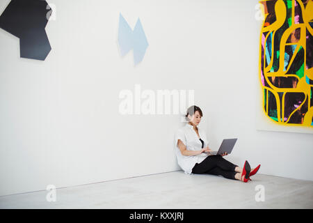 Donna con corti capelli neri che indossa una camicia bianca e pantaloni neri e rosso tacco alto scarpe seduto sul pavimento i art gallery, tenendo il laptop. Foto Stock