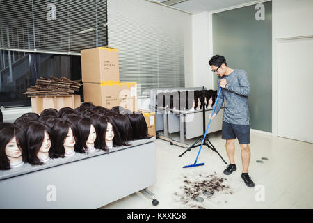 Uomo Barbuto con gli occhiali in piedi in ambienti chiusi, spazzando via i capelli sul pavimento, grande gruppo di teste di manichino marrone con parrucche sulle tabelle. Foto Stock