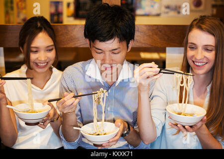 Tre persone sorridenti sidy seduta a fianco a un tavolo in un ristorante, mangiare da bocce utilizzando bastoncini. Foto Stock