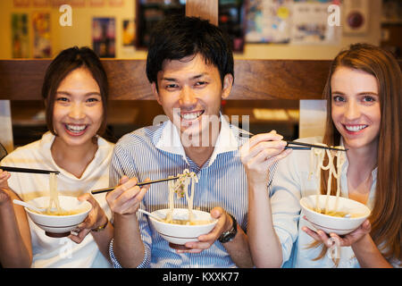 Tre persone sorridenti sidy seduta a fianco a un tavolo in un ristorante, mangiare da bocce utilizzando bastoncini. Foto Stock