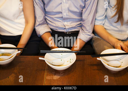 Angolo alto close up di tre persone sedute sidy a fianco a un tavolo in un ristorante, vuoto ciotole con bacchette di fronte a loro. Foto Stock