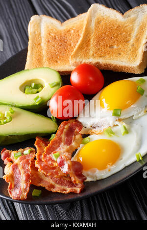 In casa le uova fritte con pancetta, cipolla, avocado, toast e pomodori close-up su una piastra nera sul piano verticale. Foto Stock