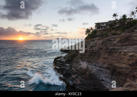 Grotte di sputare al tramonto in Hawaii Kai, Oahu con onde che si infrangono contro una scogliera sormontata con palme e case Foto Stock