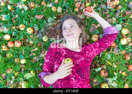 Sorridente giovane donna faccia ragazza giacente su un terreno con molti mele caduto wild fresco su erba contuso a terra su apple picking farm closeup Foto Stock