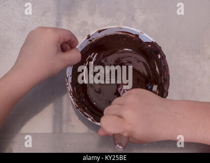 Le mani di un bambino gioca il pasticcere mescolando il cioccolato in un recipiente prima di riporlo nel cioccolatini stampi. Vista da sopra. Foto Stock