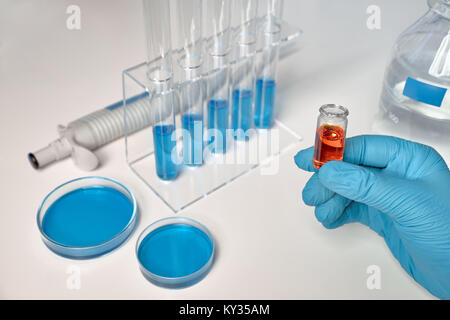 Background scientifico in blu, bianco e arancione. Il campione di liquido in mano femmina, blu campioni liquidi in tubi di vetro e piatti. Foto Stock