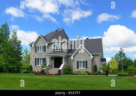 Grande casa a due piani con rivestimenti in pietra e tetto nero con cielo blu, puffy nuvole bianche, erba verde di alberi decidui e alcuni rosso dei fiori e delle piante Foto Stock